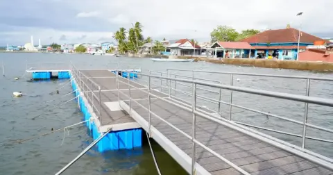 Dermaga apung panjang dengan railing dan ponton biru di perairan pesisir dekat permukiman